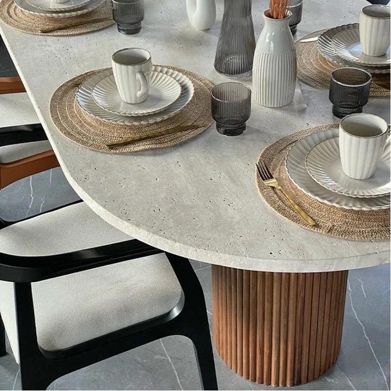 May include: A modern dining table setting with white plates, mugs, and glasses. The table is round with a light gray surface and a brown wooden base. Woven placemats and gold forks complete the elegant arrangement. The chairs have black frames and white cushions.