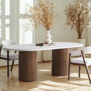 May include: An oval-shaped dining table with a white top and two cylindrical, ribbed, brown wooden supports. Two chairs with white upholstered seats and dark wooden frames are positioned on either side. A vase with dried plants sits on the table.