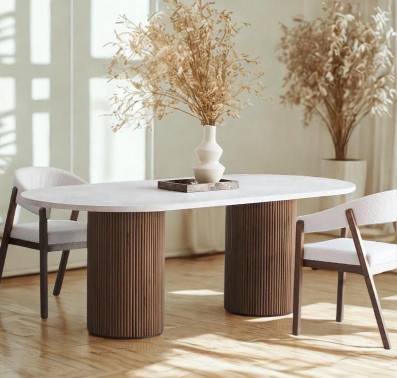 May include: An oval-shaped dining table with a white top and two cylindrical, ribbed, brown wooden supports. Two chairs with white upholstered seats and dark wooden frames are positioned on either side. A vase with dried plants sits on the table.