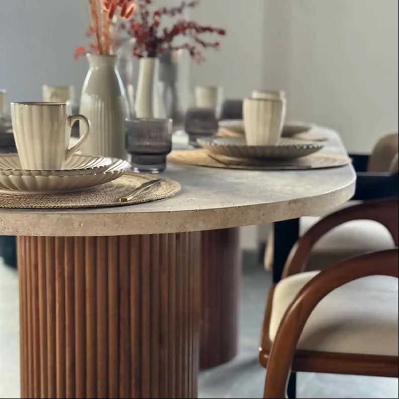 May include: A dining table set for a meal. The table has a light-colored, oval-shaped top and a cylindrical base made of vertical wooden slats. Beige plates, cups, and glasses are set on the table, along with decorative vases and flowers. The chairs have wooden frames and cream-colored cushions.