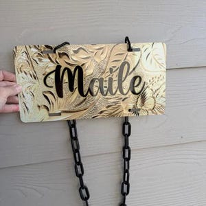 May include: A rectangular gold-toned license plate with an embossed floral pattern and the name Maile written in black script lettering. The plate features four mounting slots and is suspended by a black plastic chain link attachment.