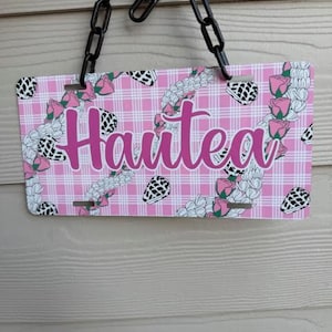 May include: A pink license plate with the word "Haute" in a cursive font. The plate has a pink plaid background with floral and black and white patterned accents. A black chain hangs from the top.