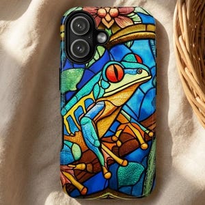 May include: A phone case featuring a stained-glass design of a red-eyed tree frog. The case has a vibrant color palette of blues, greens, yellows, and oranges. The frog is perched on a branch, and the background is a mosaic of colors.