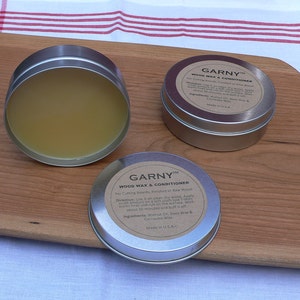 May include: Two silver tins of wood wax and conditioner, labeled "GARNY&trade; WOOD WAX & CONDITIONER". The label on the tin on the left says "For Cutting Boards, Finished or Raw Wood. Direction: Use it on clean dry wood. Apply small amount on a soft cloth (old T-shirt works fine) and rub on the surface. Wait about 30 minutes and buff it off. Ingredients: Walnut Oil, Bees Wax & Carnauba Wax. Made in U.S.A."