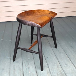 May include: A wooden stool with a curved, brown seat and black painted legs. The stool has a simple, rustic design.