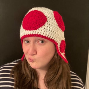 May include: A white crocheted hat with red mushroom-shaped accents. The hat has ear flaps and a playful design.