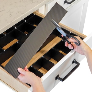 May include: A drawer organizer system with black dividers and a gray corrugated divider being cut with scissors. The drawer is open, revealing a wooden interior and white cabinet exterior with black handles. The organizer is designed to customize drawer space.