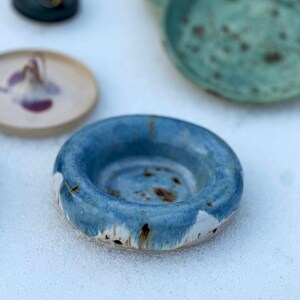 May include: A collection of handmade ceramic pieces. The foreground features a blue bowl with white and brown accents. Other items include small plates in shades of blue, green, and cream.