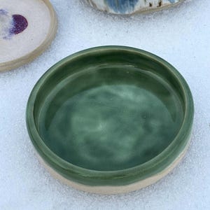 May include: A handmade ceramic bowl with a deep green interior and a light beige rim. The bowl is round and has a smooth, glossy finish. Other ceramic pieces are visible in the background.
