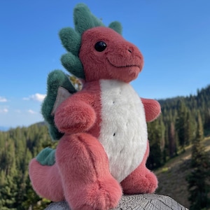 K&ouml;nnte beinhalten: Ein rotes und wei&szlig;es Pl&uuml;schdrachenspielzeug mit gr&uuml;nen Akzenten, vor dem Hintergrund einer Berglandschaft und eines blauen Himmels. Der Drache hat einen freundlichen Ausdruck.