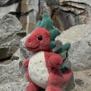 K&ouml;nnte beinhalten: Ein Pl&uuml;schdrache mit rotem K&ouml;rper, gr&uuml;nen Akzenten und einem wei&szlig;en Bauch mit schwarzen Punkten. Der Drache hat schwarze Augen und sitzt auf einem Felsen. Ein bezaubernder, handgefertigter Artikel.
