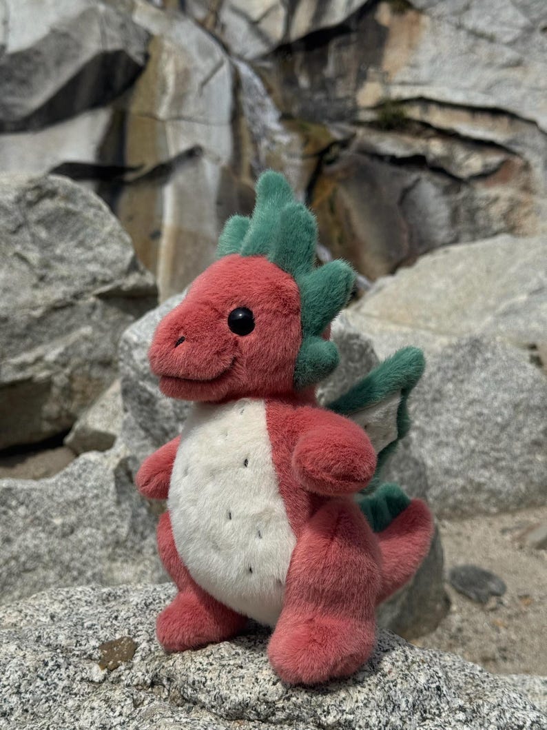 K&ouml;nnte beinhalten: Ein Pl&uuml;schdrache mit rotem K&ouml;rper, gr&uuml;nen Akzenten und einem wei&szlig;en Bauch mit schwarzen Punkten. Der Drache hat schwarze Augen und sitzt auf einem Felsen. Ein bezaubernder, handgefertigter Artikel.