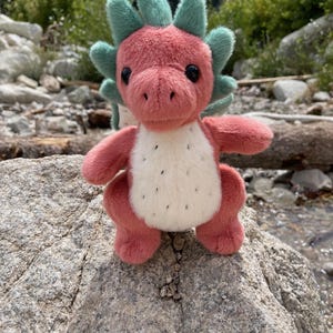 K&ouml;nnte beinhalten: Ein rosa Pl&uuml;schdrache mit wei&szlig;em Bauch und schwarzen Punkten sowie gr&uuml;nen Stacheln. Der Drache sitzt auf einem grauen Felsen, vor einem nat&uuml;rlichen Hintergrund aus Felsen, B&auml;umen und Wasser.