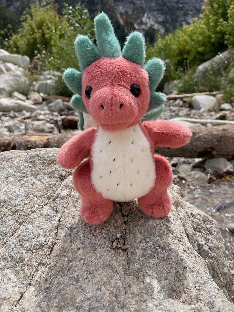 K&ouml;nnte beinhalten: Ein rosa Pl&uuml;schdrache mit wei&szlig;em Bauch und schwarzen Punkten sowie gr&uuml;nen Stacheln. Der Drache sitzt auf einem grauen Felsen, vor einem nat&uuml;rlichen Hintergrund aus Felsen, B&auml;umen und Wasser.