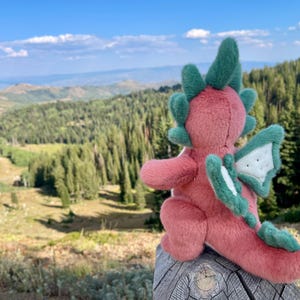 K&ouml;nnte beinhalten: Ein rosa und gr&uuml;nes Pl&uuml;schdrachenspielzeug mit gr&uuml;nen Details an Fl&uuml;geln, H&ouml;rnern und Schwanz sitzt auf einem Holzpfosten. Der Hintergrund zeigt eine malerische Aussicht auf Berge und B&auml;ume unter blauem Himmel.