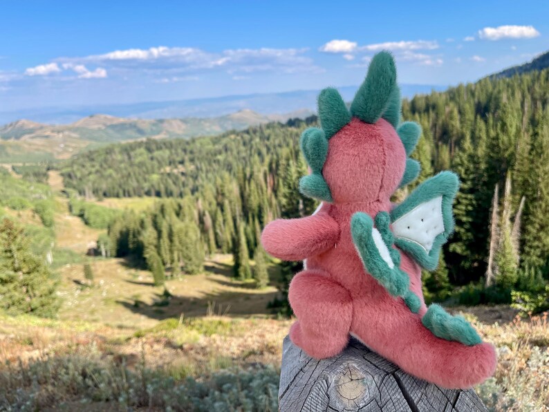 K&ouml;nnte beinhalten: Ein rosa und gr&uuml;nes Pl&uuml;schdrachenspielzeug mit gr&uuml;nen Details an Fl&uuml;geln, H&ouml;rnern und Schwanz sitzt auf einem Holzpfosten. Der Hintergrund zeigt eine malerische Aussicht auf Berge und B&auml;ume unter blauem Himmel.