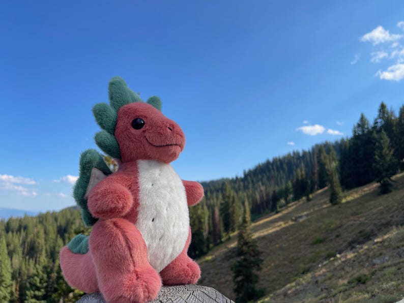 K&ouml;nnte beinhalten: Ein rotes und wei&szlig;es Pl&uuml;schdrachenspielzeug mit gr&uuml;nen Akzenten, vor dem Hintergrund einer Berglandschaft und eines blauen Himmels. Der Drache hat einen freundlichen Ausdruck.