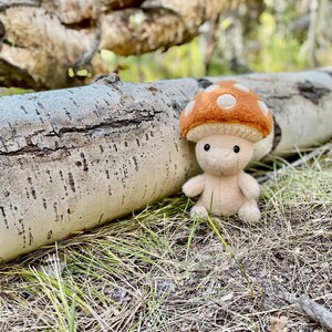 Op de afbeelding: Een vilten paddestoel knuffel met een beige lichaam en een bruine hoed met witte stippen. Het speelgoed staat op de grond in de buurt van een gevallen boomstam. De achtergrond is wazig, wat wijst op een natuurlijke buitenomgeving.