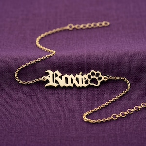May include: A gold-coloured metal bracelet featuring a central nameplate with the name Roxie in an old English font, followed by a small paw print cutout. The nameplate is attached to a delicate link chain with a lobster clasp at the end.