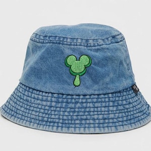 Puede incluir: Sombrero de pescador de mezclilla azul con un diseño de personaje de dibujos animados verde. El sombrero presenta una silueta clásica de sombrero de pescador con un ala estructurada y una textura de mezclilla suave y descolorida. El personaje de dibujos animados verde está bordado en la parte delantera.