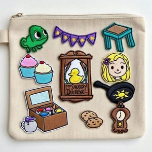 Op de afbeelding: Beige etui met ritssluiting met diverse geborduurde patches. Patches zijn onder meer een groene kameleon, cupcakes, een paarse banner, een kleine blauwe tafel, een omlijst eendenbord, een blond meisje, een koekenpan, een doos met verf, koekjes en een klok.