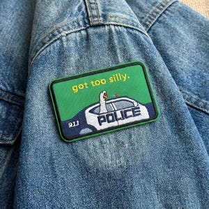 May include: A close-up of a blue denim jacket with a green patch. The patch shows a police car with a duck inside and the words "got too silly." The word "POLICE" is also visible on the patch.