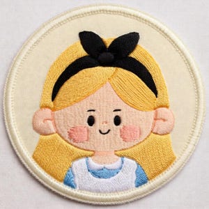 May include: Circular embroidered patch depicting a cartoon girl with blonde hair, a black bow, and a blue and white dress. The patch has a cream-coloured background and a stitched border.