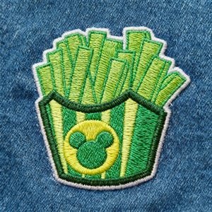 Peut inclure: &Eacute;cusson brod&eacute; repr&eacute;sentant des frites vertes et jaunes dans un contenant. Le contenant a des rayures vertes et jaunes et un cercle jaune avec une t&ecirc;te de Mickey Mouse. L'&eacute;cusson est bord&eacute; de blanc.