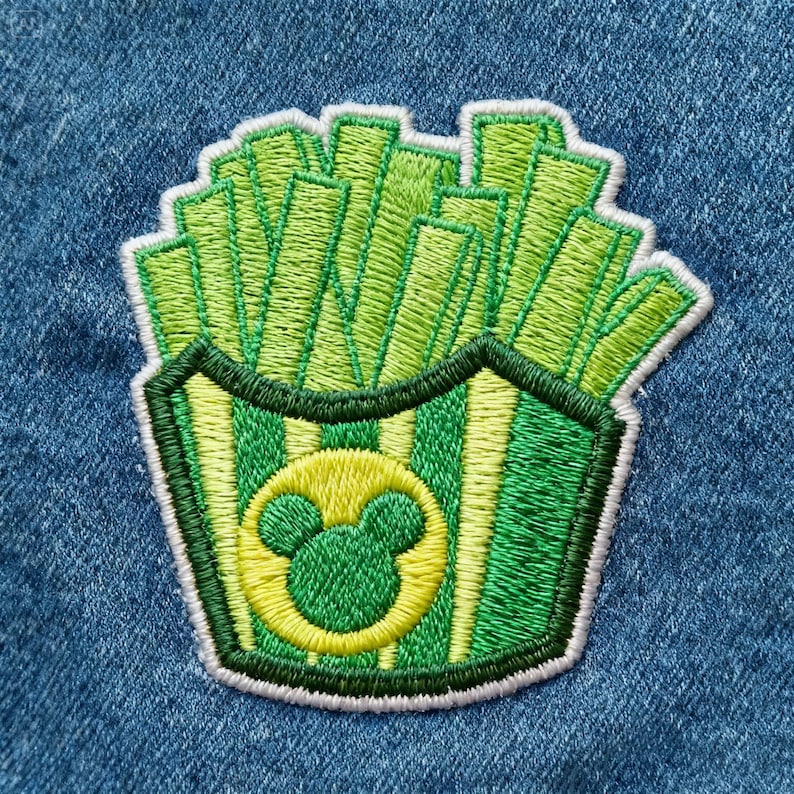 Peut inclure: &Eacute;cusson brod&eacute; repr&eacute;sentant des frites vertes et jaunes dans un contenant. Le contenant a des rayures vertes et jaunes et un cercle jaune avec une t&ecirc;te de Mickey Mouse. L'&eacute;cusson est bord&eacute; de blanc.