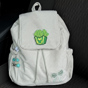 Peut inclure: Un sac &agrave; dos gris clair avec une fermeture &agrave; rabat, un cordon de serrage et des poches lat&eacute;rales. Le rabat pr&eacute;sente un &eacute;cusson vert et jaune de frites avec une t&ecirc;te de Mickey Mouse. Un n&oelig;ud et un porte-cl&eacute;s avec des perles et une &eacute;toile sont &eacute;galement visibles.
