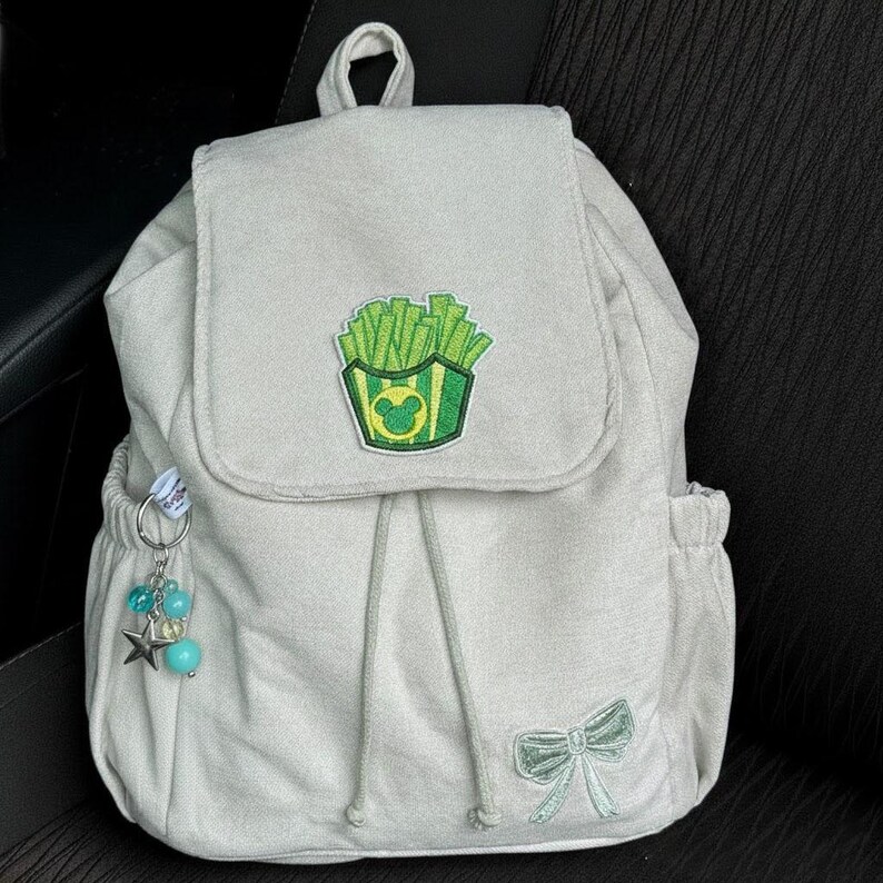 Peut inclure: Un sac &agrave; dos gris clair avec une fermeture &agrave; rabat, un cordon de serrage et des poches lat&eacute;rales. Le rabat pr&eacute;sente un &eacute;cusson vert et jaune de frites avec une t&ecirc;te de Mickey Mouse. Un n&oelig;ud et un porte-cl&eacute;s avec des perles et une &eacute;toile sont &eacute;galement visibles.