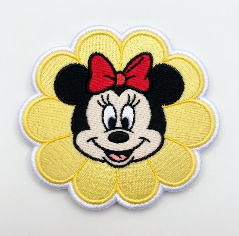 Op de afbeelding: Geborduurde patch met Minnie Mouse in het midden van een gele bloem met witte rand. Minnie heeft zwarte oren, een rode strik en een glimlach. De patch is bedoeld voor kleding of accessoires.