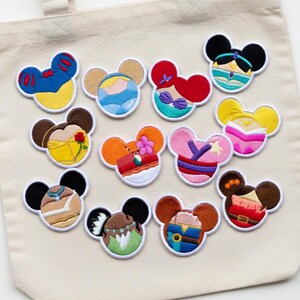 Peut inclure: Collection de patchs brodés en forme d'oreilles de Mickey Mouse, chacun représentant une princesse ou un personnage Disney différent. Les patchs sont sur un sac fourre-tout beige.