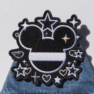 Puede incluir: Parche bordado en blanco y negro con la imagen de la cabeza de Mickey Mouse coronada, rodeada de estrellas, corazones y cruces. El parche est&aacute; sobre un fondo de mezclilla, ideal para ropa o accesorios.