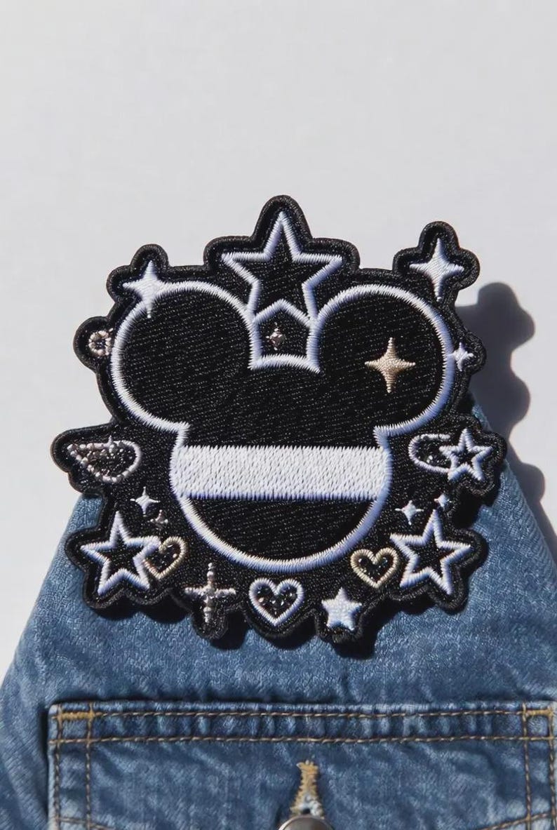 Puede incluir: Parche bordado en blanco y negro con la imagen de la cabeza de Mickey Mouse coronada, rodeada de estrellas, corazones y cruces. El parche est&aacute; sobre un fondo de mezclilla, ideal para ropa o accesorios.