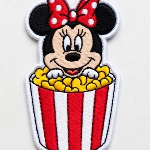 Op de afbeelding: Geborduurde patch met Minnie Mouse in een popcornemmer. Minnie draagt een rode strik met witte stippen. De popcornemmer is rood en wit gestreept, gevuld met gele popcorn.