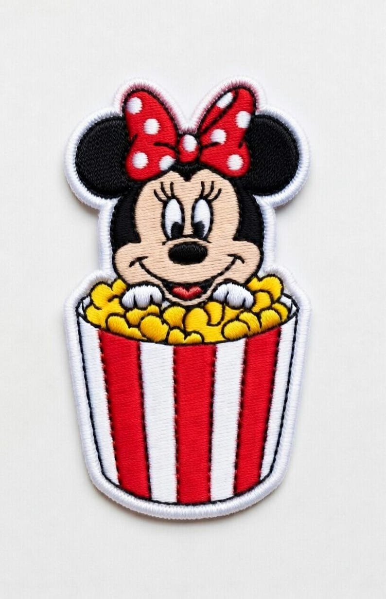 Op de afbeelding: Geborduurde patch met Minnie Mouse in een popcornemmer. Minnie draagt een rode strik met witte stippen. De popcornemmer is rood en wit gestreept, gevuld met gele popcorn.