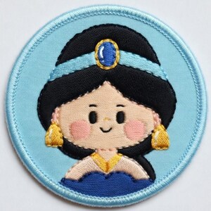 May include: A circular embroidered patch depicting a cartoon princess with black hair, a blue headband, and gold earrings. The princess has rosy cheeks and a smiling face. The patch is set against a light blue background with a blue border.