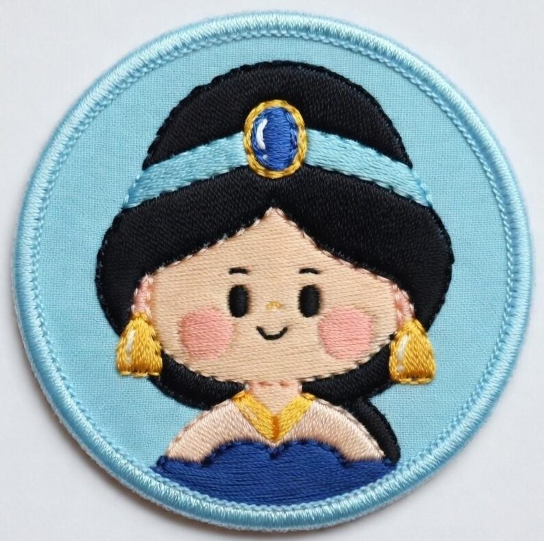 May include: A circular embroidered patch depicting a cartoon princess with black hair, a blue headband, and gold earrings. The princess has rosy cheeks and a smiling face. The patch is set against a light blue background with a blue border.