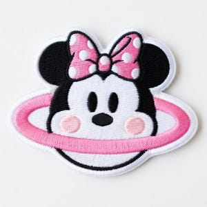 Op de afbeelding: Geborduurde patch met een cartoonmuis met een roze strik en een roze ring rond het gezicht. De muis heeft zwarte oren, ogen en neus, met roze wangen. De patch is wit met zwarte en roze accenten.