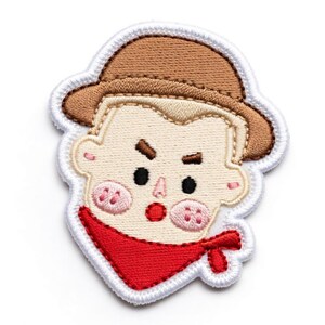 Puede incluir: Parche bordado con la cara de un vaquero de dibujos animados. El dise&ntilde;o incluye un sombrero marr&oacute;n, un pa&ntilde;uelo rojo y mejillas rosadas. El parche est&aacute; contorneado con costuras blancas.