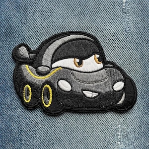 Puede incluir: Parche bordado con un coche de dibujos animados con una cara sonriente. El coche es principalmente negro con detalles amarillos en las ruedas y una raya amarilla. Los ojos del coche son blancos con iris marrones. El parche est&aacute; sobre un fondo de mezclilla azul.