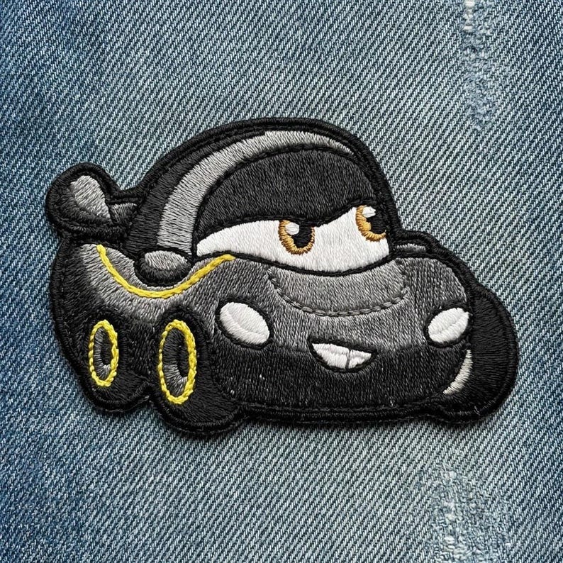 Puede incluir: Parche bordado con un coche de dibujos animados con una cara sonriente. El coche es principalmente negro con detalles amarillos en las ruedas y una raya amarilla. Los ojos del coche son blancos con iris marrones. El parche est&aacute; sobre un fondo de mezclilla azul.