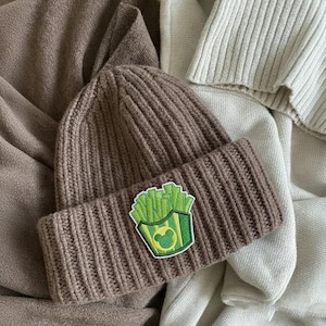 Peut inclure: Un bonnet en tricot c&ocirc;tel&eacute; marron avec un bord repli&eacute;. Le bonnet pr&eacute;sente un &eacute;cusson vert et jaune d'un contenant de frites avec un motif de t&ecirc;te de Mickey Mouse. Le chapeau est pr&eacute;sent&eacute; sur un fond neutre.