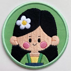 May include: Embroidered patch depicting a cartoon girl with black hair, pink cheeks, and a white flower. She wears a green and yellow outfit, set against a light green circular background with a green border. Ideal for clothing or accessories, this patch is a decorative item.