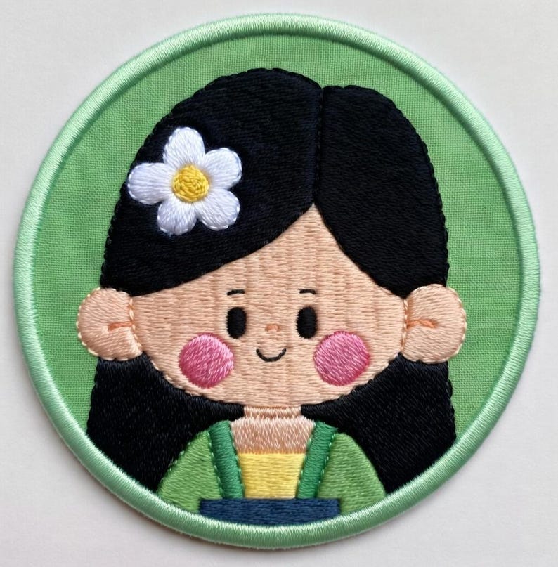 May include: Embroidered patch depicting a cartoon girl with black hair, pink cheeks, and a white flower. She wears a green and yellow outfit, set against a light green circular background with a green border. Ideal for clothing or accessories, this patch is a decorative item.