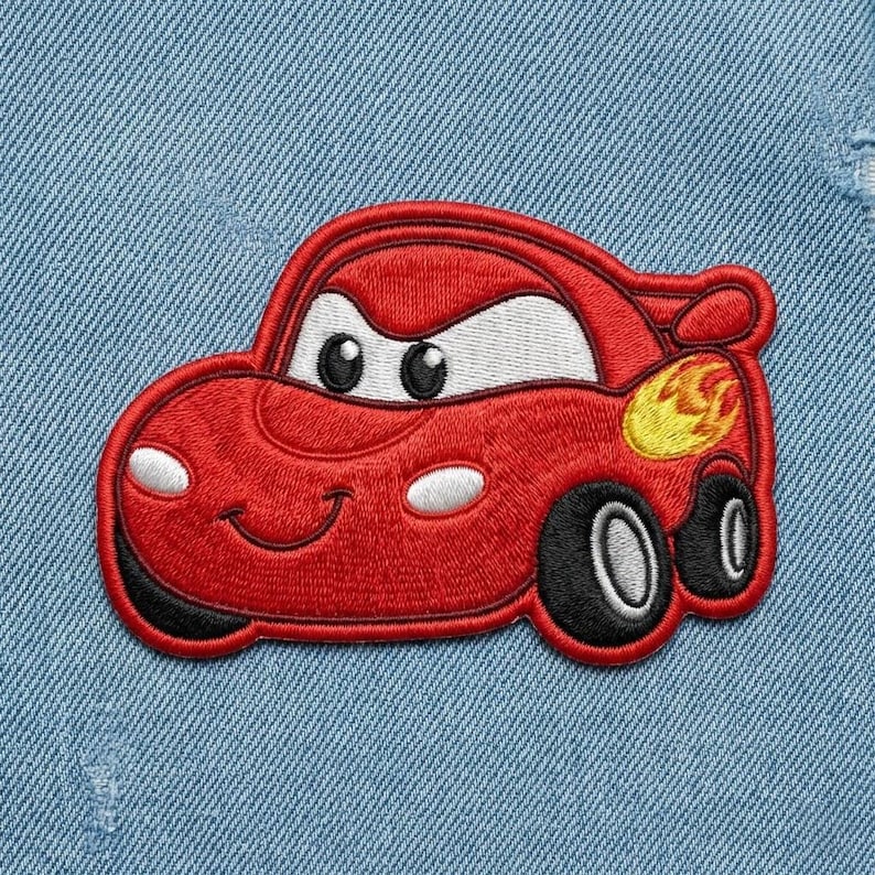 Puede incluir: Un parche de coche rojo bordado con una cara sonriente, ojos blancos y neum&aacute;ticos negros. El parche tiene un dise&ntilde;o de llama amarilla y naranja en el lateral. El parche est&aacute; sobre un fondo de mezclilla azul claro.