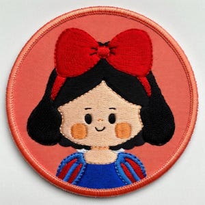 May include: Embroidered patch with a cartoon illustration of a character with black hair, a red bow, and a blue dress. The patch has a red border and a peach-coloured background. The design is a circular shape.