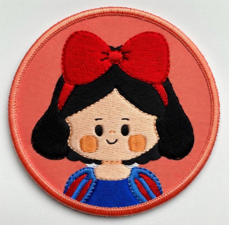 May include: Embroidered patch with a cartoon illustration of a character with black hair, a red bow, and a blue dress. The patch has a red border and a peach-coloured background. The design is a circular shape.