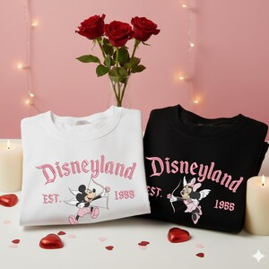 May include: Two sweatshirts, one white and one black, with the word "Disneyland" in pink script. The white sweatshirt features Mickey Mouse as Cupid, and the black one features Minnie Mouse. Both have "EST. 1955" below the word Disneyland.